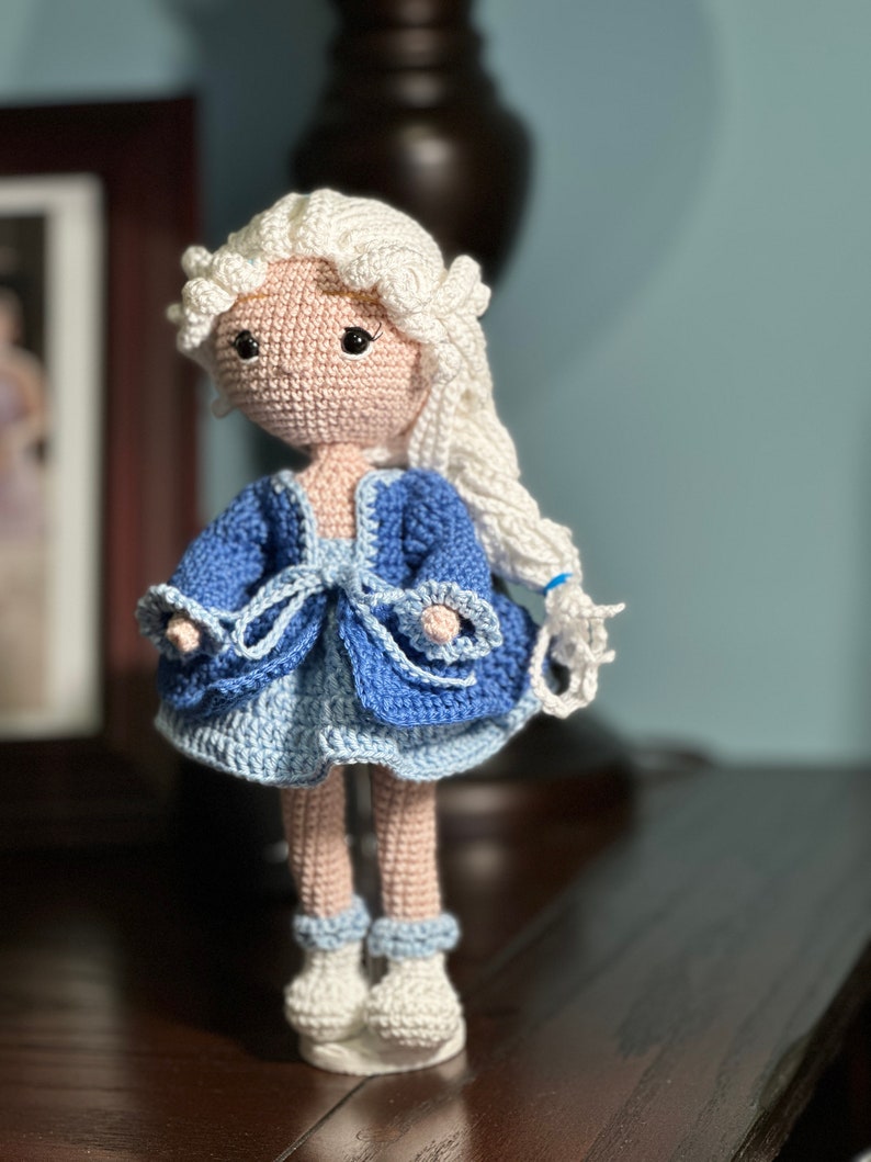 May include: A handmade crochet doll with a light skin tone, white hair, and black eyes. The doll wears a blue dress with a light blue skirt and white shoes. The doll is standing on a white base.