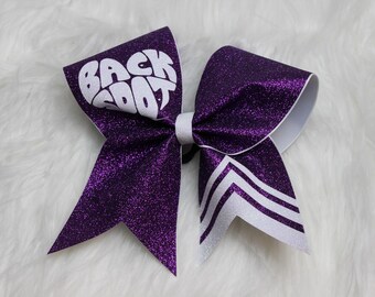 May include: A purple glitter cheer bow with the text "BACK SCOOT" in white. The bow has white accents and a white center knot. The bow is on a white fluffy surface.