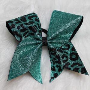 May include: A teal glitter cheer bow with black leopard print accents. The bow is made of a shiny material and has a black elastic band. The bow is a hair accessory.