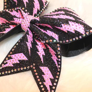 May include: Close-up of two black glitter bows with pink lightning bolt designs. The bows are embellished with rhinestones around the edges and center. The bows are on a white fluffy surface.