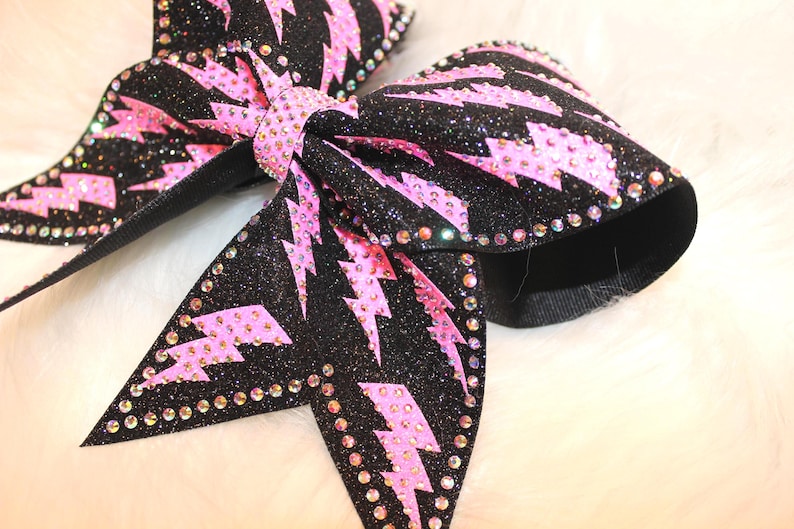 May include: Close-up of two black glitter bows with pink lightning bolt designs. The bows are embellished with rhinestones around the edges and center. The bows are on a white fluffy surface.