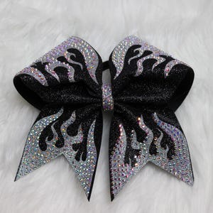 Op de afbeelding: Zwarte cheerleading strik met een glitterafwerking en een vlammenontwerp omlijnd met zilveren strass. De strik is gecentreerd met een bijpassende strassband.