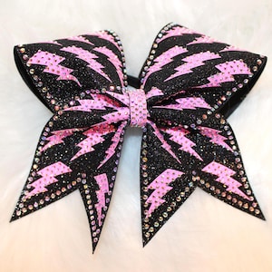 May include: A large, black and pink cheerleading bow with a sparkling glitter finish. The bow features pink lightning bolt designs and is embellished with rhinestones along the edges and center. This accessory is perfect for cheerleaders.