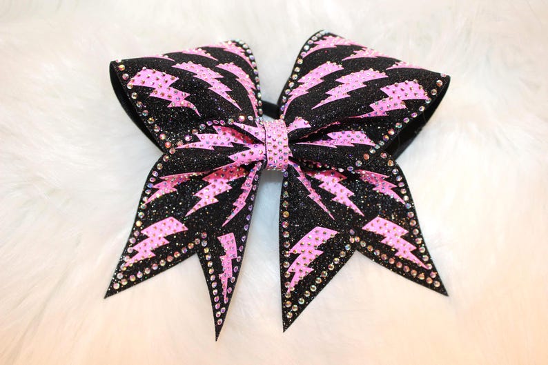 May include: A large, black and pink cheerleading bow with a sparkling glitter finish. The bow features pink lightning bolt designs and is embellished with rhinestones along the edges and center. This accessory is perfect for cheerleaders.