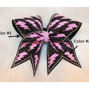 May include: A large, glittery black and pink cheerleading bow. The bow features a pink lightning bolt pattern and is embellished with rhinestones along the edges. The center of the bow is also pink.