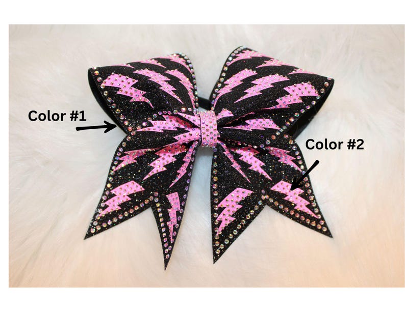 May include: A large, glittery black and pink cheerleading bow. The bow features a pink lightning bolt pattern and is embellished with rhinestones along the edges. The center of the bow is also pink.