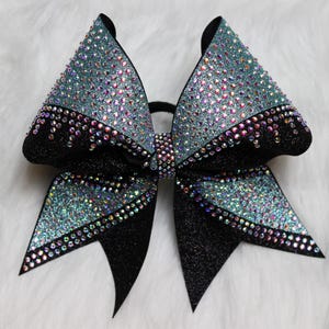 Op de afbeelding: Een grote, glinsterende cheerleading strik met een zwarte basis en teal accenten. De strik is rijkelijk versierd met iriserende strass-steentjes, wat een sprankelend effect creëert. De strik is ontworpen met een klassieke strikvorm en een zwarte elastische band.