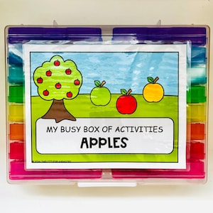 May include: A clear plastic storage container with colorful compartments, featuring a label titled MY BUSY BOX OF ACTIVITIES APPLES. The label shows a green apple tree, a green apple, a red apple, and a yellow apple on a grassy field.