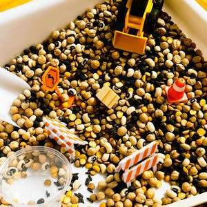 May include: A sensory bin filled with a mixture of dried black-eyed peas, chickpeas, and black beans. The bin contains a yellow toy construction vehicle, orange traffic cones, miniature road signs, white plastic barriers, and a clear cup.