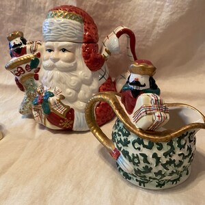 May include: A ceramic Santa Claus teapot and creamer set. The teapot is shaped like Santa Claus with a red hat and white beard. The creamer is shaped like a sack with a green and gold design.