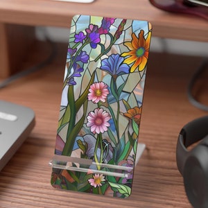 May include: A colorful stained glass phone stand with a floral design. The stand features a variety of flowers, including pink, purple, blue, and yellow. The stand is made of glass and has a clear, glossy finish.