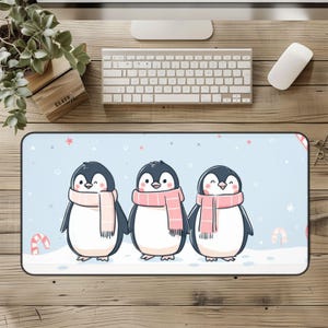 May include: A rectangular desk mat featuring three cartoon penguins wearing pink scarves, standing on a snowy surface against a light blue background. The mat has a black border and includes candy cane graphics.