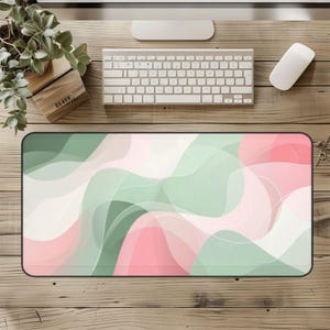 May include: A rectangular desk mat with a black border featuring an abstract design of flowing shapes in shades of pink, green, and white. A keyboard, mouse, and other office supplies are visible on a wooden desk.