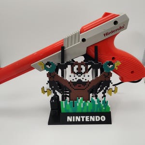 Op de afbeelding: Een rode en grijze Nintendo Zapper gun rust op een zwarte standaard met een gepixeld eendenjacht-tafereel. De standaard heeft het woord "NINTENDO" in witte blokletters. Een klassiek videogame-accessoire.