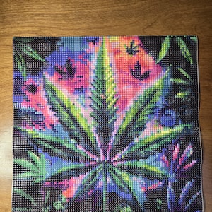 Puede incluir: Pintura de diamantes cuadrada con un diseño de hoja de cannabis. La obra de arte utiliza un estilo de mosaico con pequeños diamantes de imitación cuadrados en tonos verdes, azules, rosas y morados sobre un fondo negro. La imagen está sobre una superficie de madera.