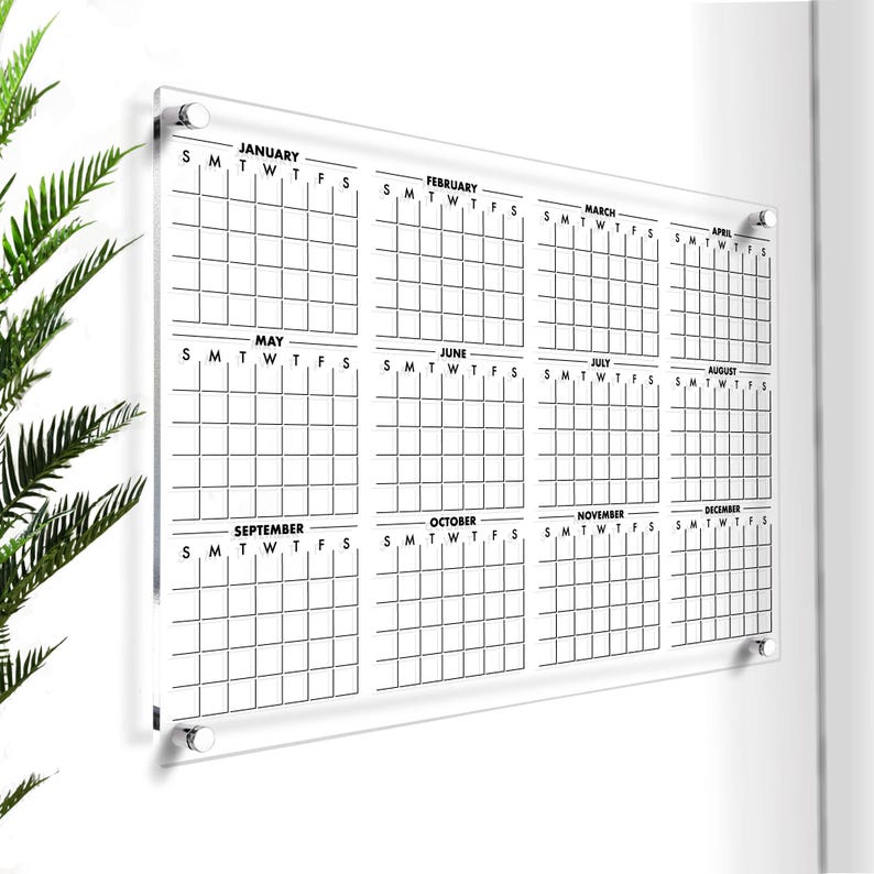 May include: A clear acrylic dry-erase calendar with a grid layout for each month of the year. The calendar includes the days of the week, S, M, T, W, T, F, S, and the months January through December.