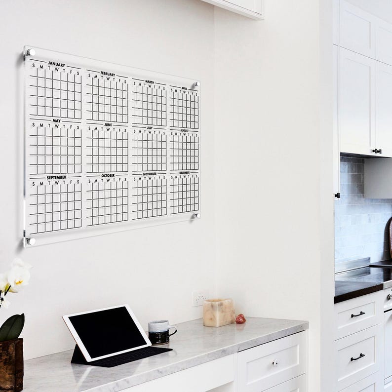 May include: A clear acrylic dry-erase calendar with black grid lines and month labels. The calendar is mounted on a white wall above a desk with a laptop, a mug, and a small decorative object.