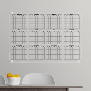 May include: A clear acrylic dry-erase calendar with a grid layout for each month of the year. The calendar is labelled with the months January through December and the days of the week, Sunday through Saturday.