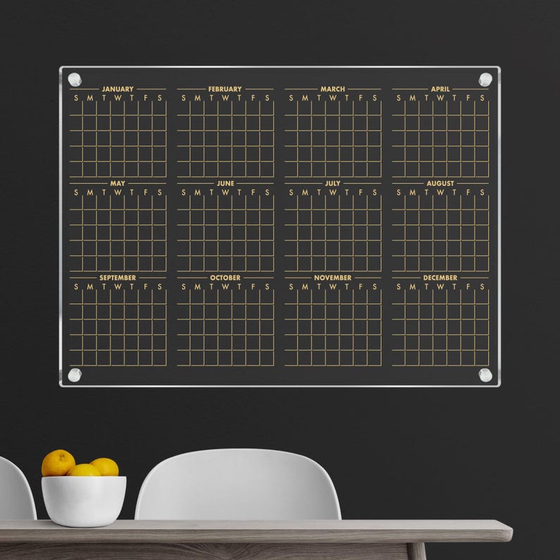 May include: A clear acrylic dry-erase calendar with gold lettering and a grid for each month of the year. The calendar is mounted on a black wall.