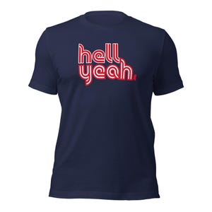 Könnte beinhalten: Ein dunkelblaues Kurzarm-T-Shirt mit dem Text "hell yeah.", der in einer stilisierten, mehrschichtigen Schriftart mit weißer Schrift und roter Umrandung gedruckt ist. Das Design ist mittig auf der Brust platziert, mit Rundhalsausschnitt.