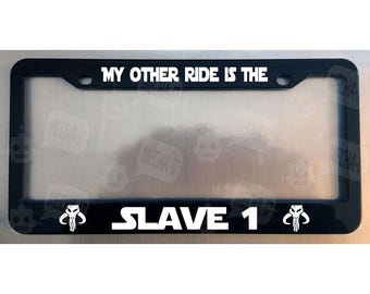 Könnte beinhalten: Ein schwarzer, rechteckiger Kennzeichenhalter mit weißem Text, der oben MY OTHER RIDE IS THE und unten SLAVE 1 liest. Die untere Leiste enthält zudem auf beiden Seiten des Textes ein weißes Symbol eines mandalorianischen Totenkopfes.