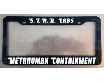 May include: A black rectangular license plate frame featuring white text. The top section displays S.T.A.R. LABS, while the bottom section reads METAHUMAN CONTAINMENT. The frame includes two circular mounting holes at the top for vehicle attachment.