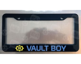 May include: A black rectangular license plate frame featuring a yellow gear icon with the number 111 inside, followed by the text VAULT BOY in light blue capital letters. The frame has two mounting holes at the top for vehicle attachment.