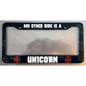May include: Black license plate frame with white text that reads "My other ride is a unicorn". The frame has two red and black Deadpool logos on either side of the text.