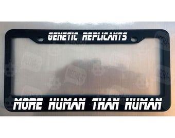 May include: A black rectangular license plate frame featuring white text. The top section displays the words GENETIC REPLICANTS, while the bottom section features the phrase MORE HUMAN THAN HUMAN in a bold, stylized, sans-serif font.