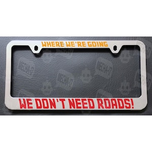 May include: A silver license plate frame with the text "Where We're Going We Don't Need Roads!" in orange and red lettering.