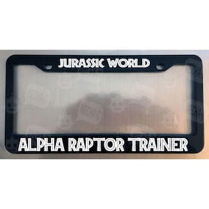 May include: Black license plate frame with white text that reads "Jurassic World Alpha Raptor Trainer".