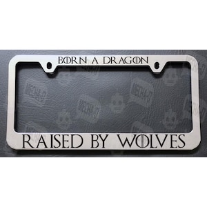 May include: A silver metal license plate frame with the text "BORN A DRAGON RAISED BY WOLVES" in black lettering.