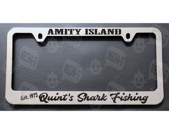 Może przedstawiać: Srebrna metalowa ramka na tablicę rejestracyjną z czarnym tekstem, który brzmi "Amity Island Est. 1975 Quint's Shark Fishing".