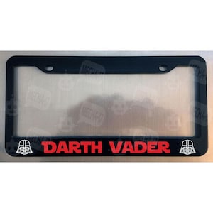 May include: Black plastic license plate frame with a red "Darth Vader" text and two white Darth Vader silhouette images on either side.
