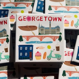 May include: A set of colorful square magnets with the word "GEORGETOWN" printed in red. Each magnet features a different illustration of a building, a boat, a cupcake, a scooter, and other details.
