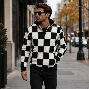 May include: A long-sleeve zip-up jacket featuring a bold black and white checkered pattern throughout. The garment includes a high collar, a central front zipper, and solid black ribbed cuffs and hem, worn over a plain black shirt and black pants.