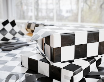 May include: Two gift boxes wrapped in black and white checkered paper are stacked on a matching surface. A roll of the same paper and a pair of scissors are visible in the background. A black ribbon is draped across the surface.