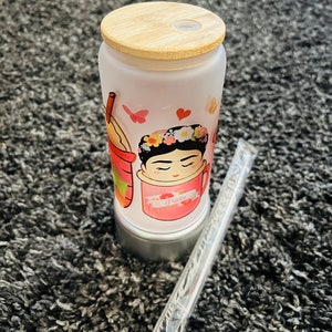 May include: A frosted glass tumbler with a bamboo lid and a colorful illustration of a woman with a flower crown. The tumbler has the word "Frida" written on it. A clear plastic straw is included.