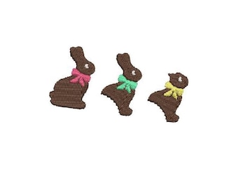 Peut inclure: Trois lapins de Pâques en chocolat avec des nœuds de différentes couleurs. Le lapin à gauche a un nœud rose, le lapin du milieu a un nœud vert et le lapin à droite a un nœud jaune.