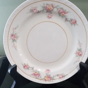 May include: A white porcelain plate with a gold rim and a floral design featuring pink and yellow roses. The plate is on a black stand.