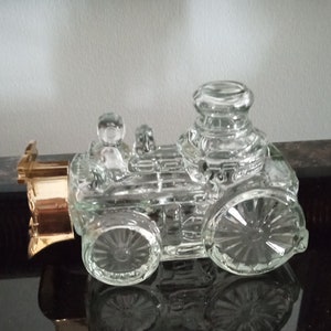 May include: A clear glass bottle shaped like a vintage train engine. The bottle has a gold-coloured cap and is sitting on a black surface.