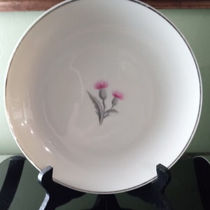 May include: White ceramic bowl with a gold rim and a pink thistle floral design. The bowl is displayed on a black stand. The bowl is a decorative piece for serving or display.
