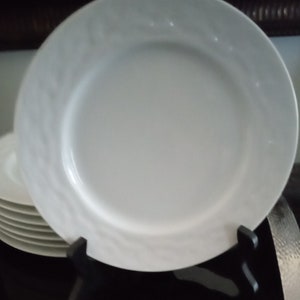 May include: A stack of white ceramic plates with a textured rim design. The plates are displayed on a black stand, with one plate in the foreground. The plates are suitable for serving meals.