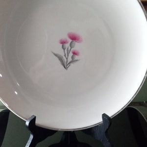 May include: A white ceramic plate with a silver rim features a delicate floral design of pink thistles on a gray stem. The plate is displayed on a black stand.