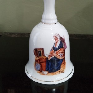 May include: White porcelain bell with a gold trim. The bell features a painting of a woman sitting in a chair, sewing a quilt. The painting is signed "Norman Rockwell".