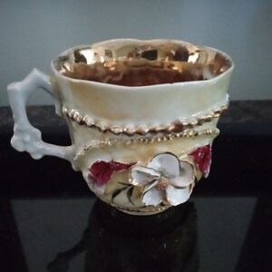 Könnte beinhalten: Eine weiße Porzellan-Teetasse mit goldfarbenem Rand und Blumenmuster. Die Tasse hat einen zarten Henkel und ist mit roten und weißen Blumen verziert.