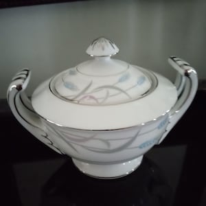 Peut inclure: Sucrier en porcelaine blanche avec un couvercle argenté et un motif floral délicat représentant des épis de blé bleus et gris. Le bol a deux poignées argentées.