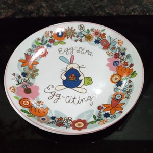 May include: A white ceramic plate with a colorful floral border and a cartoon bunny holding an egg. The text "Egg-stra Egg-citing" is written on the plate.