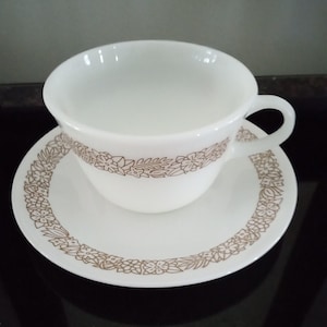 Puede incluir: Una taza de té y un platillo de cerámica blanca con un patrón floral marrón alrededor del borde.