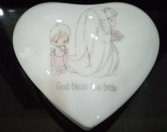 Puede incluir: Caja de cerámica blanca en forma de corazón con una imagen pintada a mano de dos niñas pequeñas. La niña de delante lleva un vestido rosa y la niña de atrás lleva un vestido blanco. El texto "God bless the bride" está impreso en la caja.